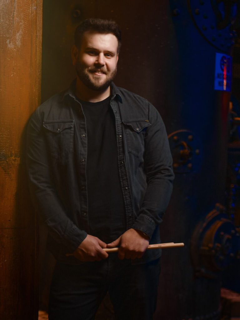 Jason Maleney is smiling and holding drumsticks, leaning against a wall.