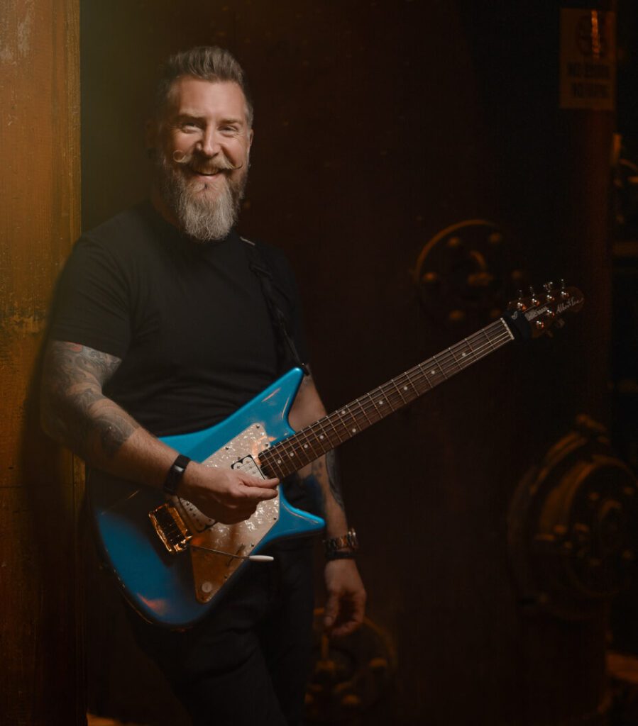 Philip Joyce is smiling and holding a blue electric guitar.